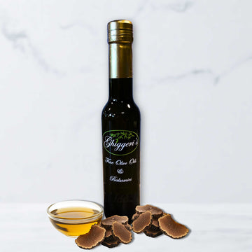 Bottle of Ghiggeri's Black Truffle Infused Olive Oil with fresh black truffle slices and a small bowl of golden oil on a marble background.