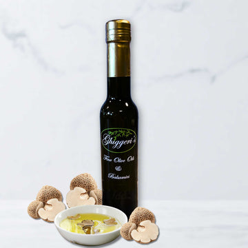 A bottle of Ghiggeri's White Truffle Infused Olive Oil alongside fresh sliced white truffles on a light marble surface.