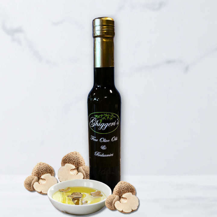 A bottle of Ghiggeri's White Truffle Infused Olive Oil alongside fresh sliced white truffles on a light marble surface.